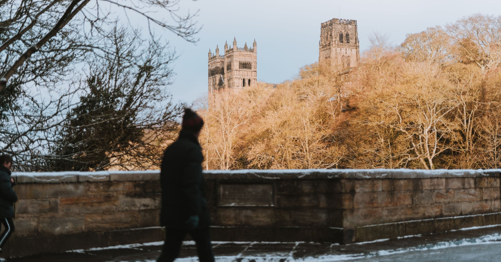 man walking across prebends bridge in durham city during winter with snow on ground and durham cathedral in background.